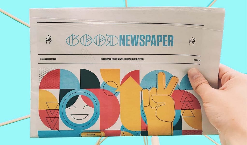 Uma mão segura um jornal chamado Good Newspaper em um fundo azul. A imagem no jornal é uma representação artística feito com formas geométricas coloridas vermelhas, azuis, amarelas e pretas. Entre elas, um rosto sorrindo e uma mão fazendo o símbolo da paz.