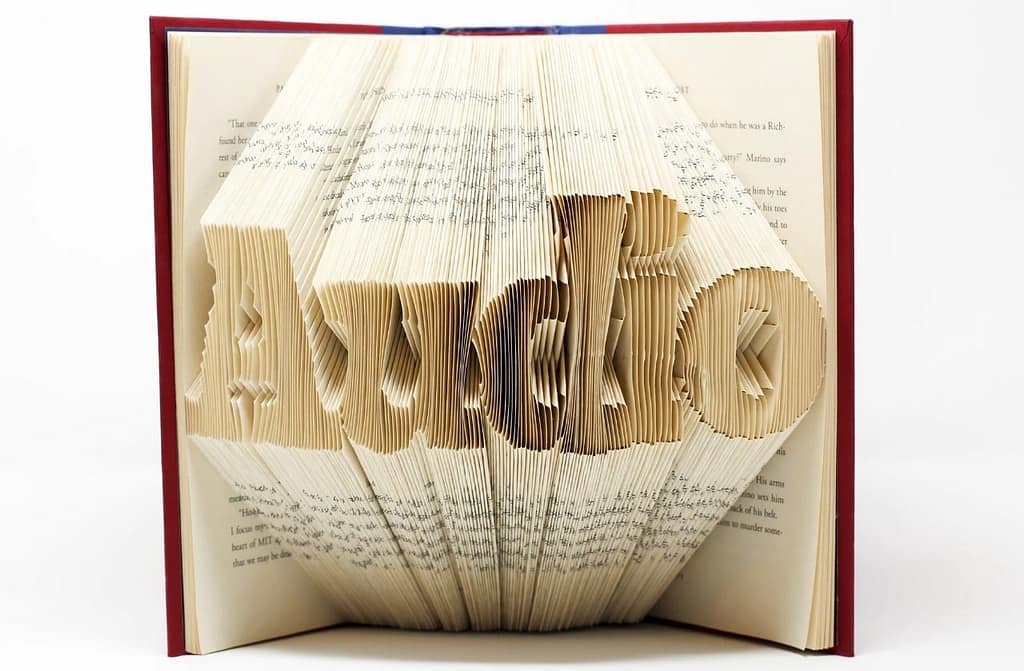Um livro aberto apoiado em pé em uma superfície branca, as páginas estão dobradas de maneira a formarem a palavra Audio.