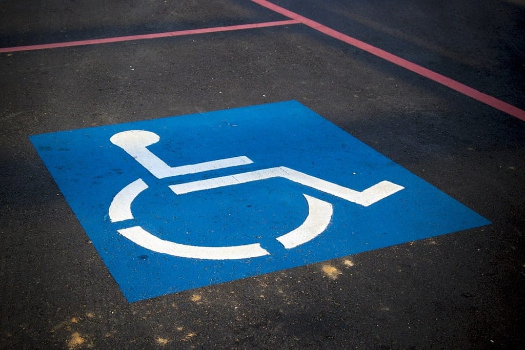 Chão de asfalto com faixas vermelhas pintadas demarcando uma vaga para carros. No meio da vaga está pintado um quadrado azul com um boneco cadeirante branco no centro, o símbolo internacional de acesso.