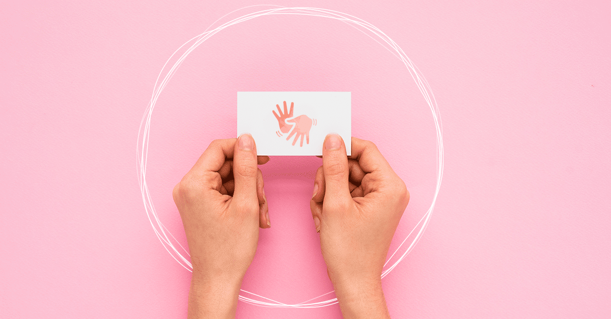 Sobre um fundo rosa claro, duas mãos seguram um cartão branco com um símbolo rosa de duas mãos abertas, uma com os dedos para cima e outra para baixo. Ao redor da imagem há um círculo branco um tracejado como se feito à lápis e reforçado por ele várias vezes.