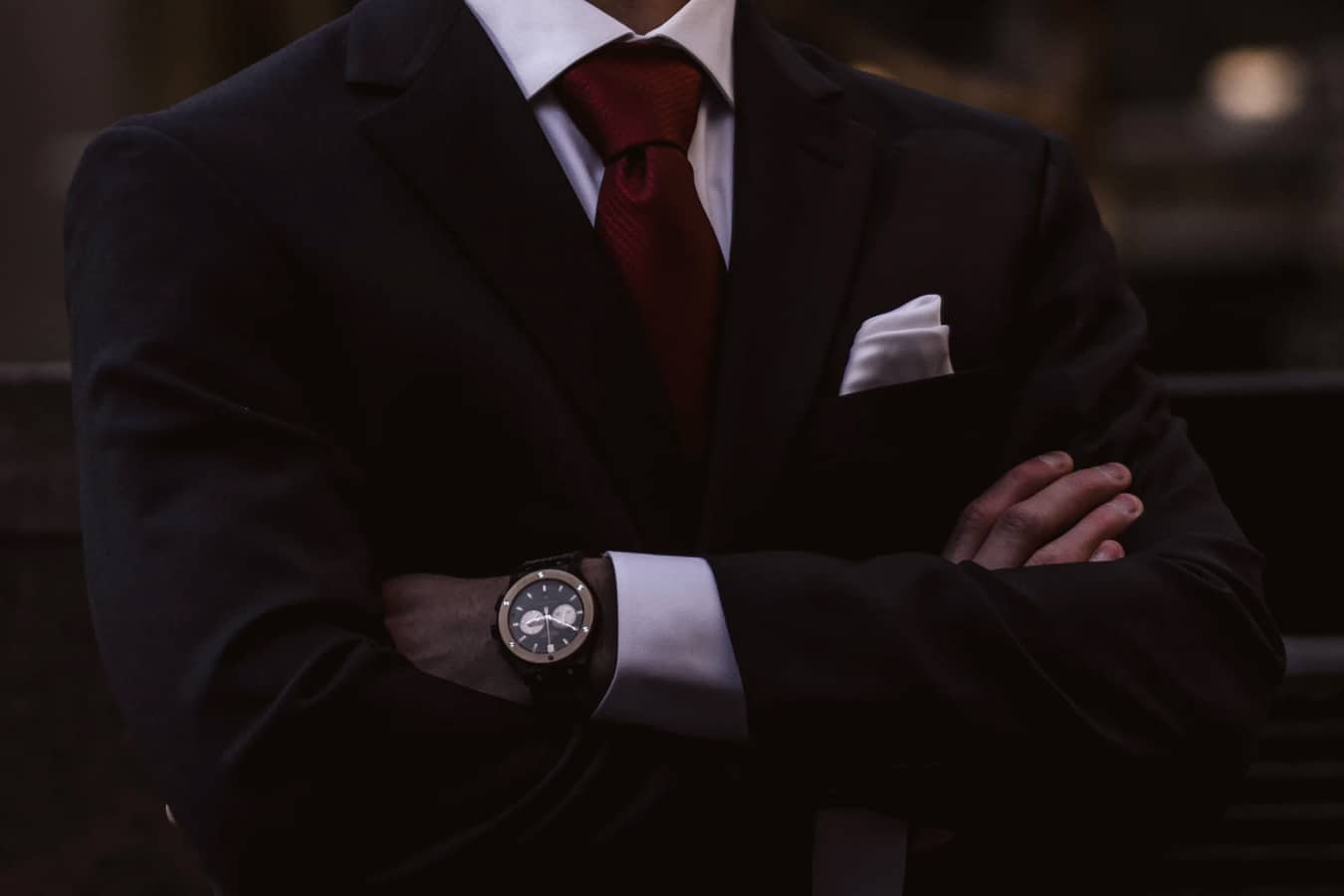 Foto do dorso de um homem de braços cruzados, ele usa um terno preto, grava vermelha e relógio no pulso.