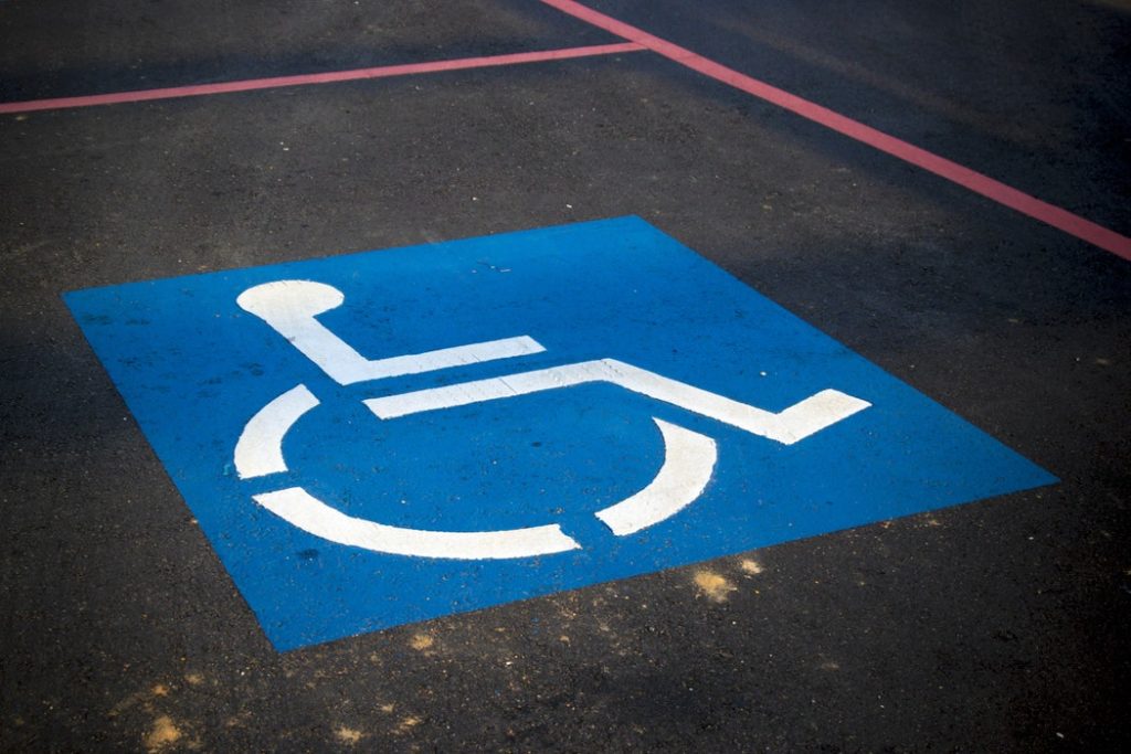 Chão de asfalto com linhas vermelhas demarcando uma vaga para carros. No chão está pintado o símbolo internacional de acesso, um quadrado azul com um boneco branco cadeirante no centro.